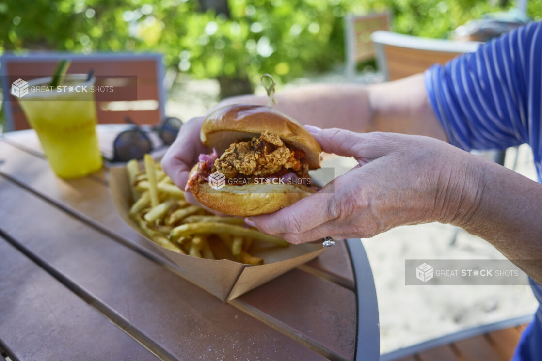 Close-Up of Hands Holding Up a Fried Chicken Burger with a Take-Out Container of French Fries on a Patio Table in an Outdoor Area of a Resort in Nassau, Bahamas