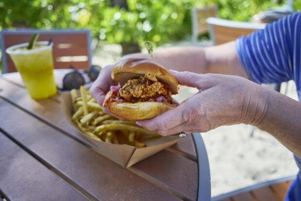 Close-Up of Hands Holding Up a Fried Chicken Burger with a Take-Out Container of French Fries on a Patio Table in an Outdoor Area of a Resort in Nassau, Bahamas