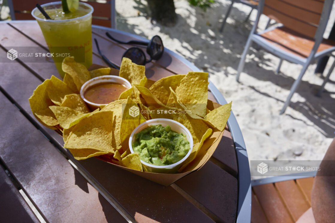 Close-Up of Patio Table with a Cardboard Container of Nacho Chips, Salsa and Guacamole and a Cold Tropical Beverage on a Sandy Beach in a Resort in Nassau, Bahamas
