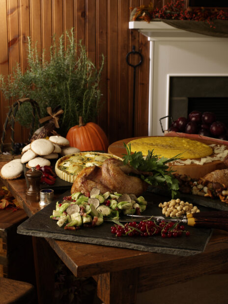 Thanksgiving Table Spread on Wooden Table with Roasted Turkey, Brussels Sprouts and Truffle Side Dish, Quiche, Breads and Cookies in Front of a Fireplace in an Indoor Setting