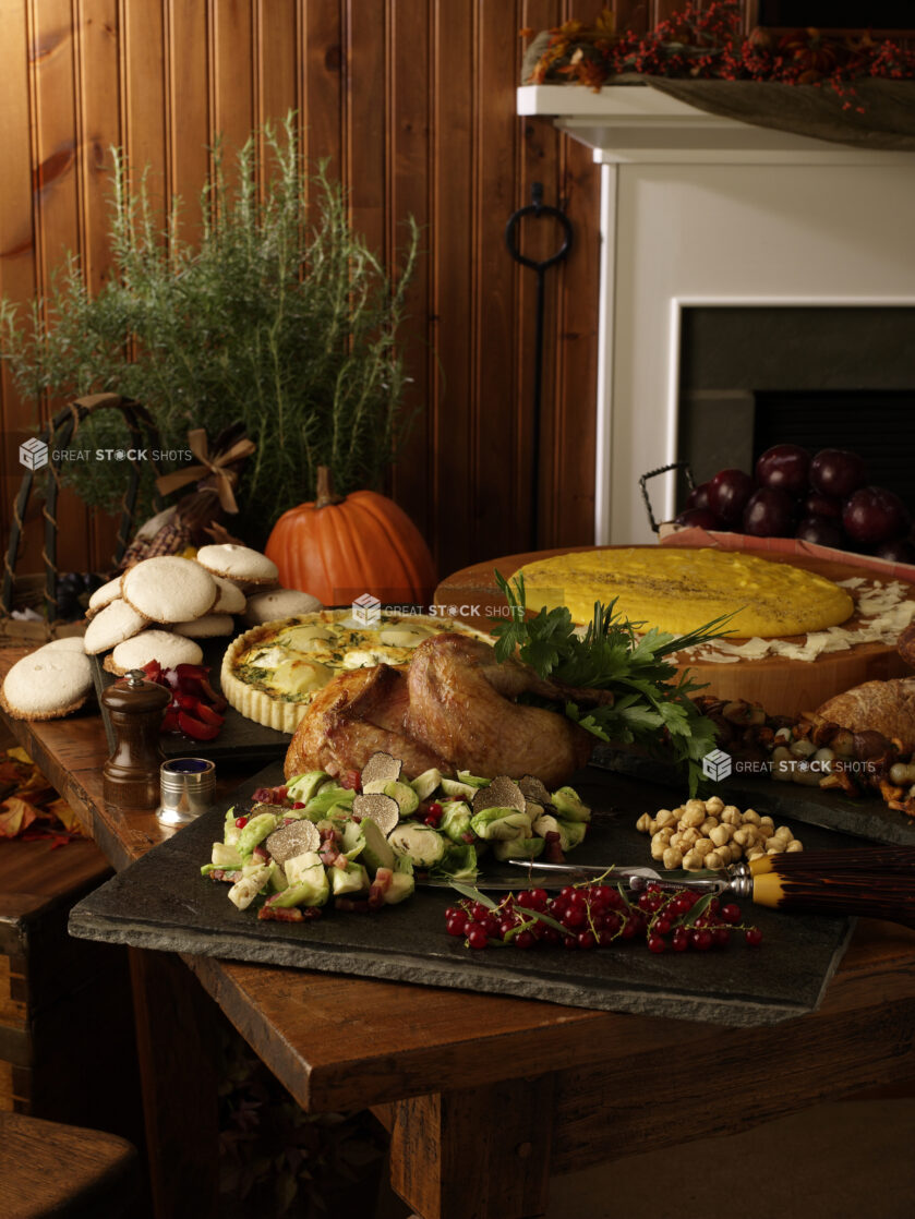 Thanksgiving Table Spread on Wooden Table with Roasted Turkey, Brussels Sprouts and Truffle Side Dish, Quiche, Breads and Cookies in Front of a Fireplace in an Indoor Setting