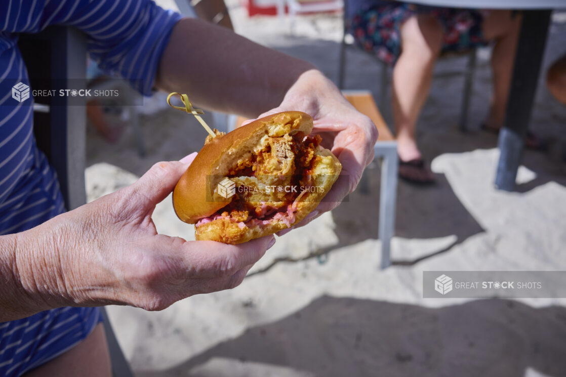 Close-Up of Hands Holding a Fried Chicken Sandwich with a Bite Taken Out of It Against a White Sand Beach Background