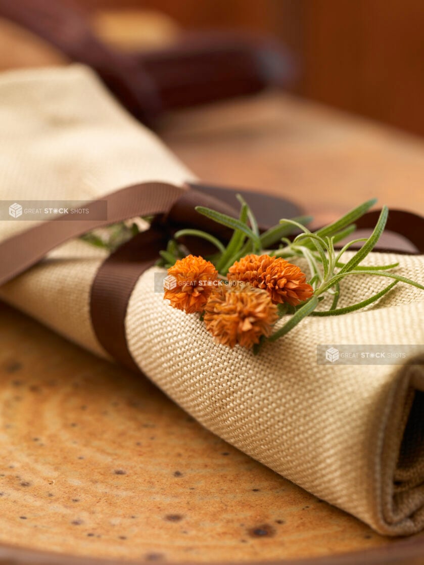 Table Setting with a Beige Placemat Rolled Up and Tied with a Dark Brown Ribbon and Orange Flowers and Fresh Leaves on a Ceramic Trivet in an Indoor Setting