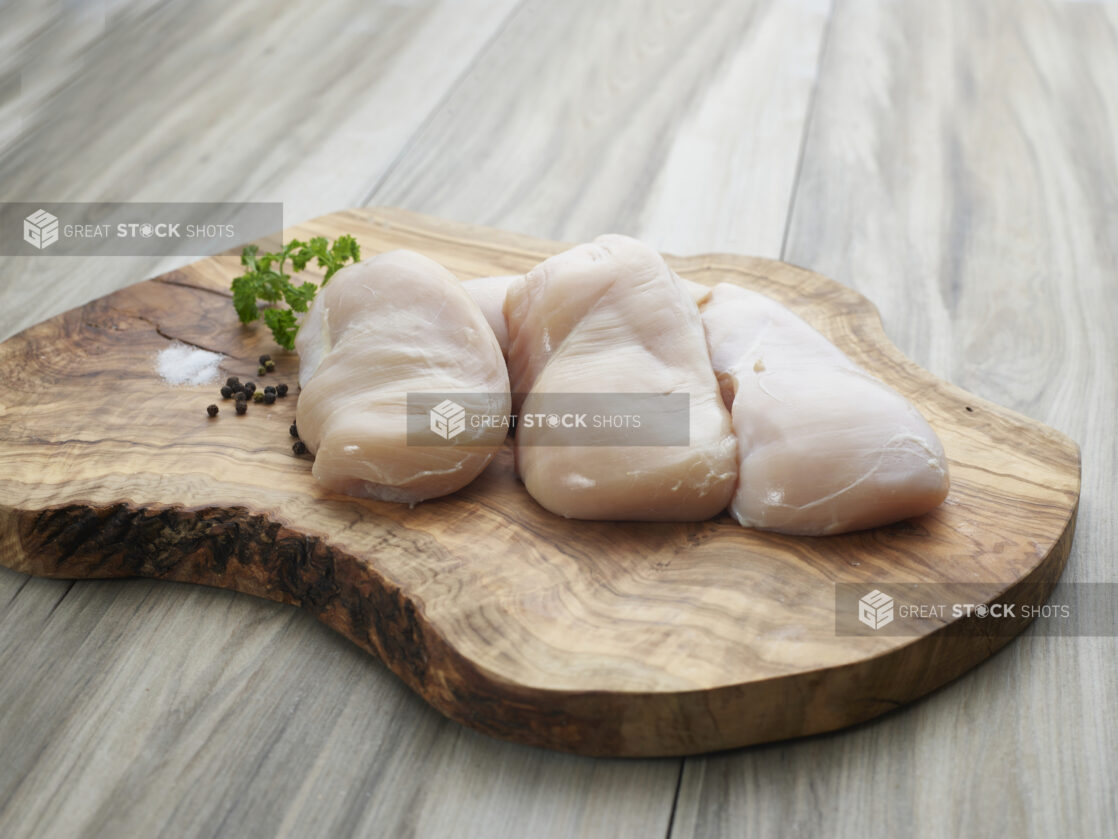 Three raw chicken breast fillets on a natural wooden board with seasonings