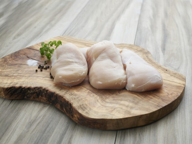 Three raw chicken breast fillets on a natural wooden board with seasonings