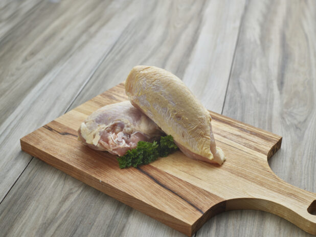 Whole raw chicken breast with skin and bones, split in two on a wood paddle, close-up