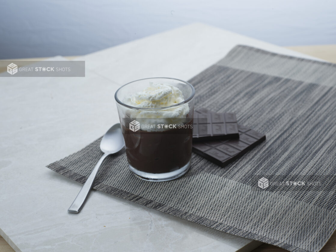 Chocolate pudding with whipped cream in a glass with a spoon, dark chocolate bar in background