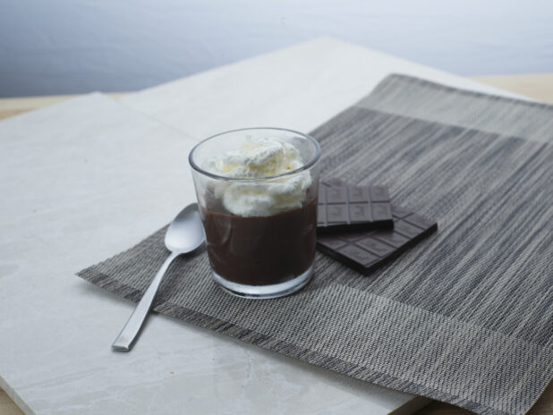 Chocolate pudding with whipped cream in a glass with a spoon, dark chocolate bar in background