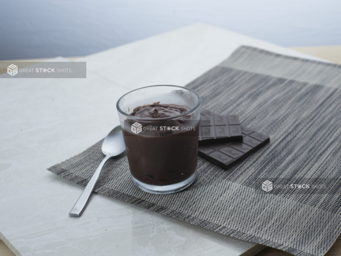 Chocolate pudding in a short glass with a spoon, dark chocolate bar in background