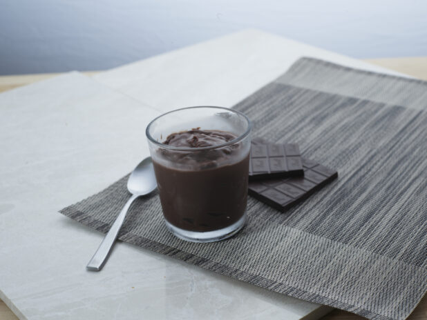 Chocolate pudding in a short glass with a spoon, dark chocolate bar in background