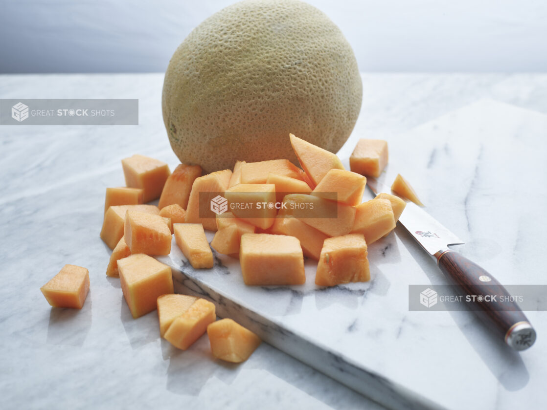 Chunks of canteloupe with knife on white marble board with whole canteloupe in background