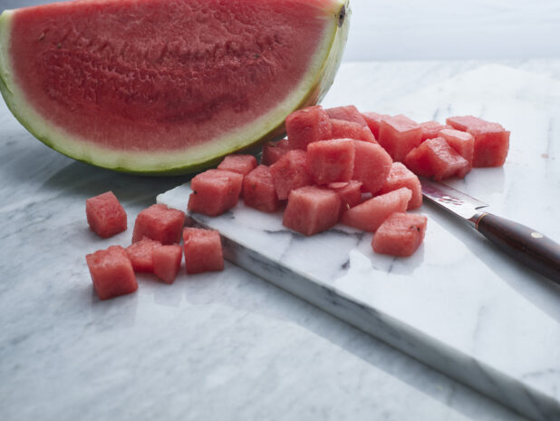Chunks of juicy watermelon with knife on white marble board with large wedge of watermelon behind