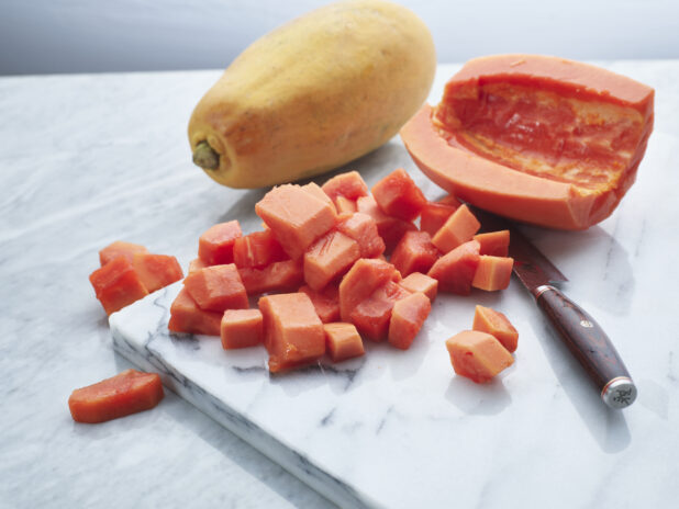 Chopped papaya with whole fruit in background on a white marble board