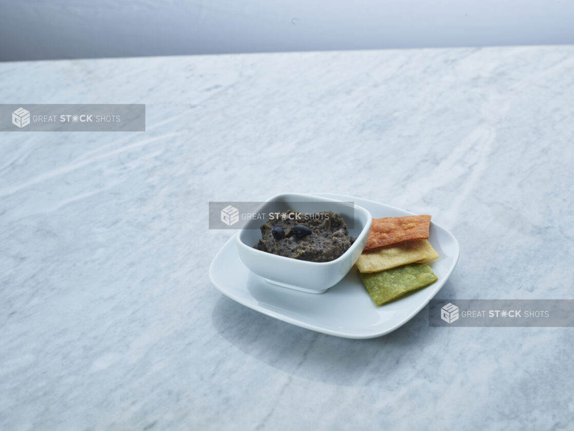 Black bean dip in a white ceramic ramekin on a plate with multicoloured tortilla chips