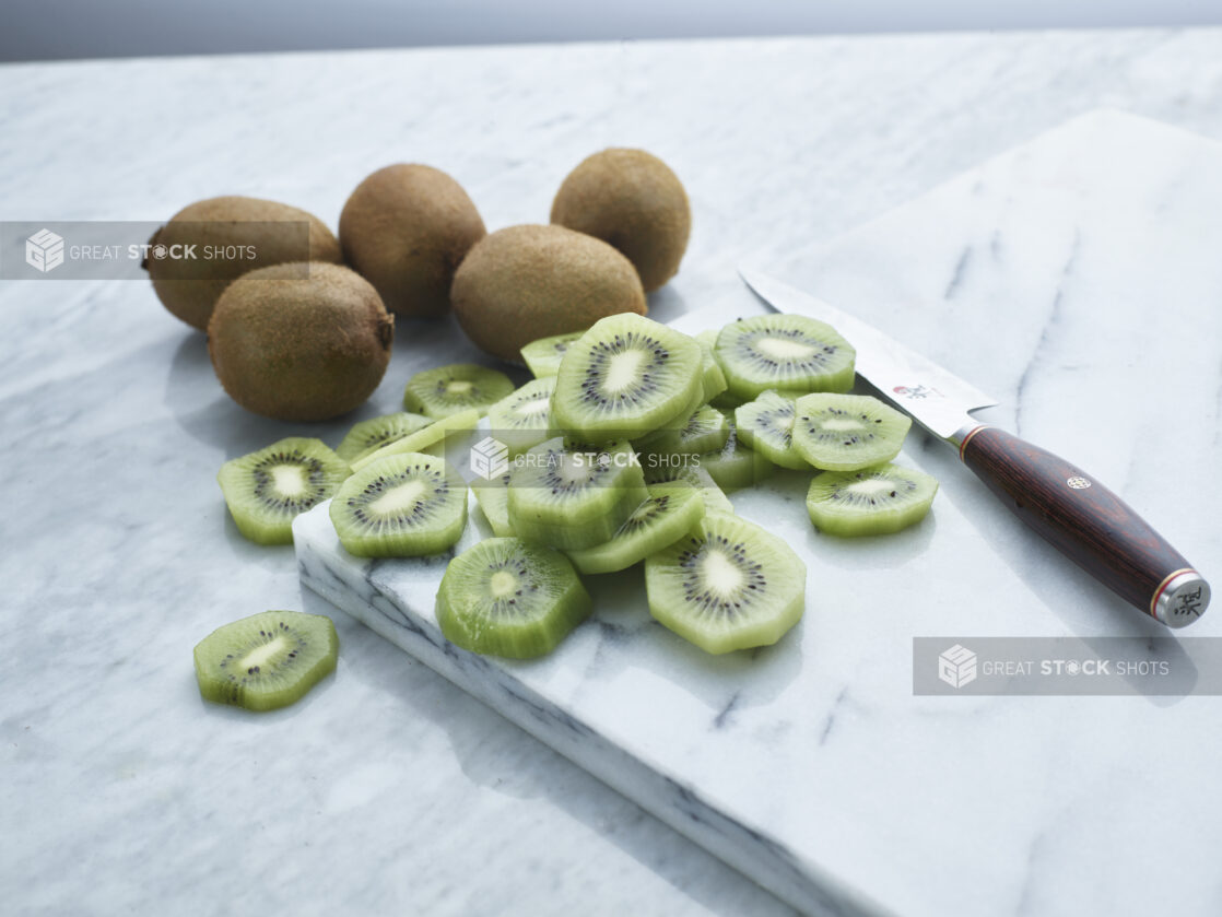 Slices of kiwi with Japanese knife on white marble board, whole kiwis in background, close-up