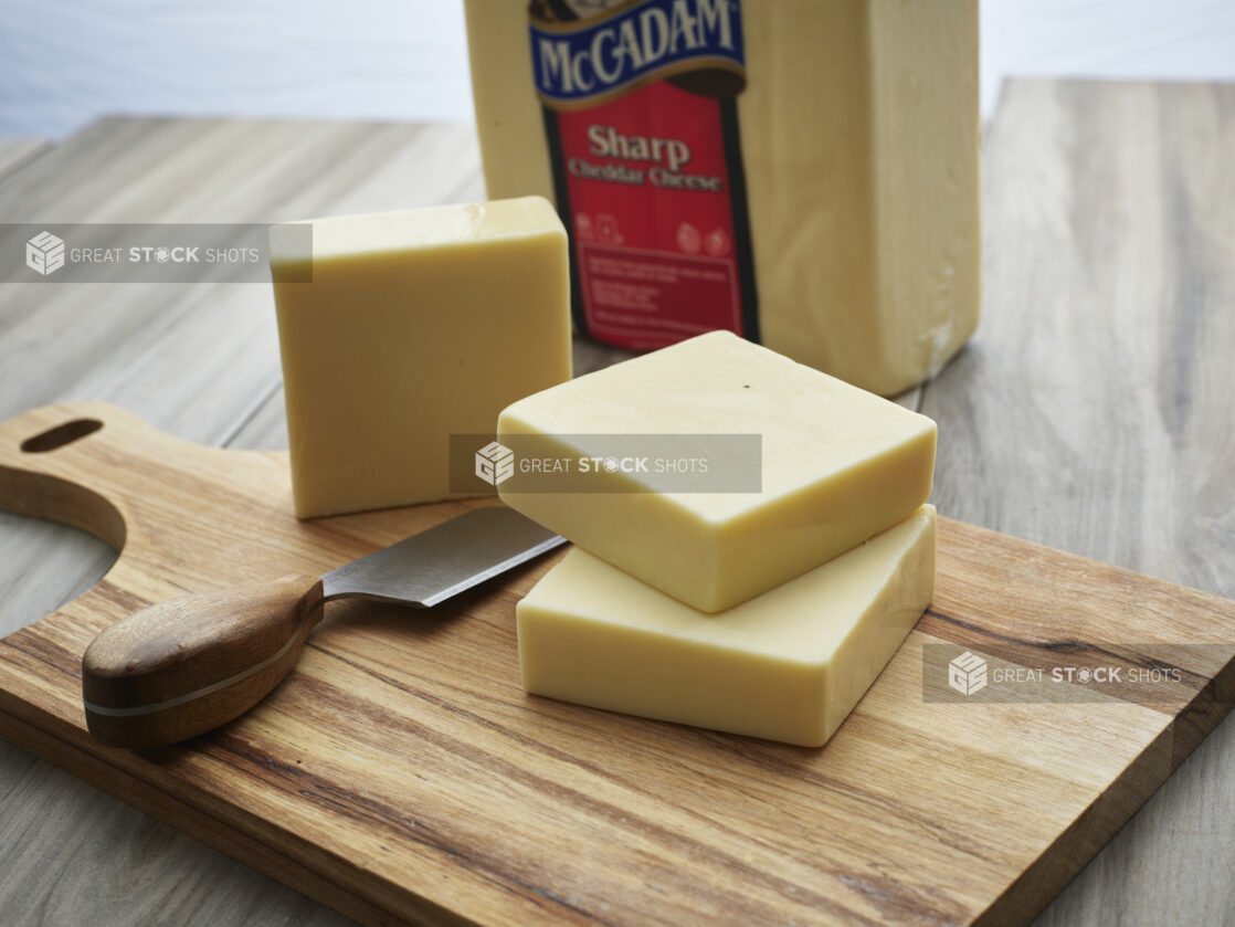 Blocks of white cheddar cheese stacked on a wooden paddle with wood-handled cheese knife, close-up