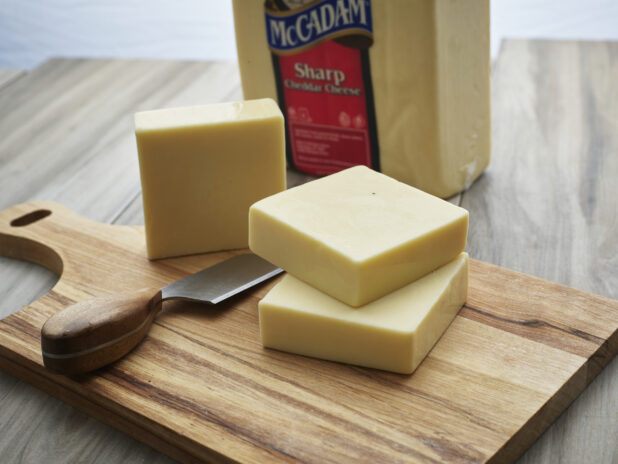 Blocks of white cheddar cheese stacked on a wooden paddle with wood-handled cheese knife, close-up