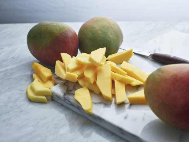 Sliced mango with whole fruit in foreground and background on a white marble board