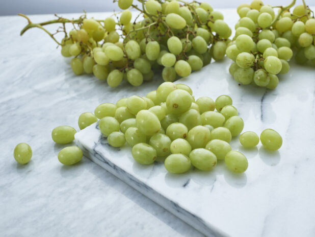 Loose green grapes on a white marble board, bunches of grapes in background, close-up