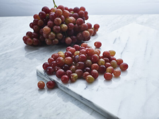 Loose red grapes on a white marble board, bunch of grapes in background, close-up