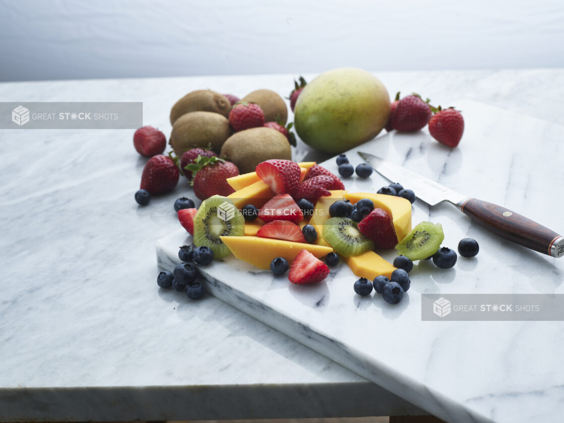 Assorted chopped fruits and berries with a Japanese knife on a white marble board, whole fruits behind