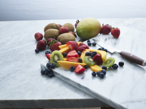 Assorted chopped fruits and berries with a Japanese knife on a white marble board, whole fruits behind