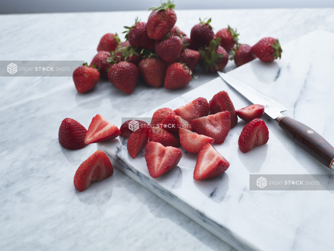 Fresh halved strawberries with knife on white marble board, whole strawberries in background, close-up