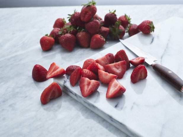 Fresh halved strawberries with knife on white marble board, whole strawberries in background, close-up