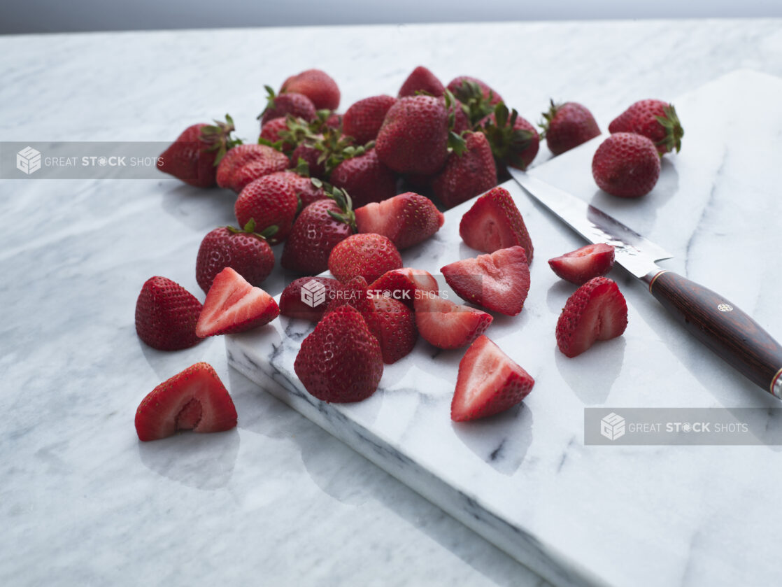 Fresh halved and whole strawberries with knife on white marble, close-up