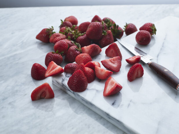 Fresh halved and whole strawberries with knife on white marble, close-up