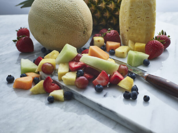 Assorted chopped fruits and berries with knife on a white marble board, close-up
