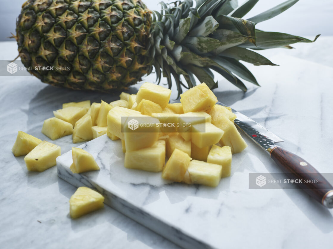 Chunks of fresh pineapple with a knife on a white marble board, whole pineapple in background