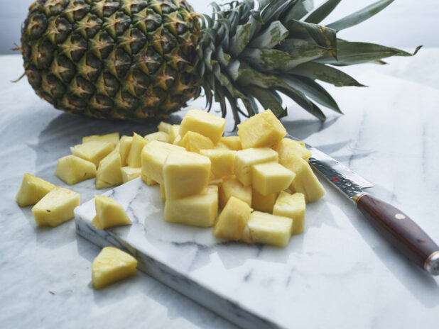 Chunks of fresh pineapple with a knife on a white marble board, whole pineapple in background