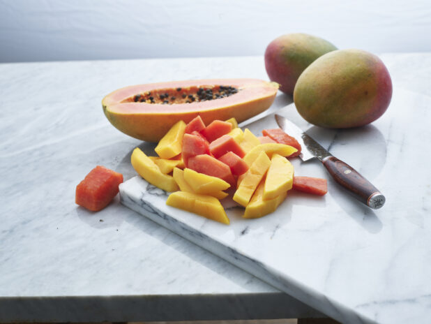 Sliced mango and papaya on with Japanese knife on white marble, close-up, whole and halved fruit in background