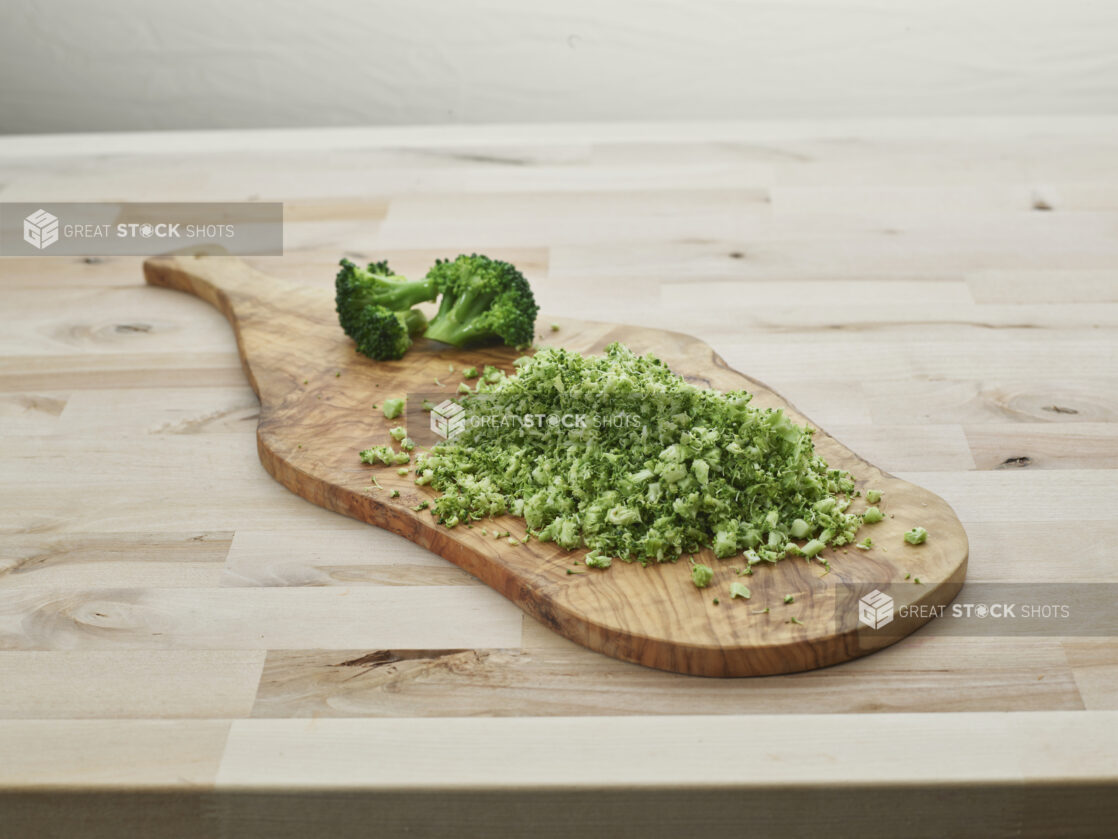 Finely chopped broccoli in a pile on a wooden paddle with broccoli florets in background