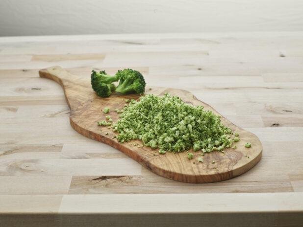 Finely chopped broccoli in a pile on a wooden paddle with broccoli florets in background