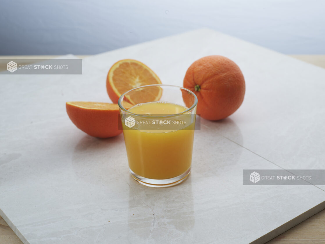 Small glass of freshly squeezed orange juice with whole and halved oranges in background, close-up