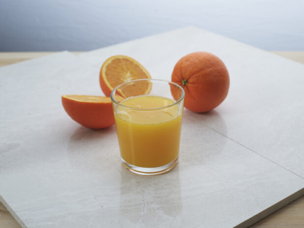 Small glass of freshly squeezed orange juice with whole and halved oranges in background, close-up