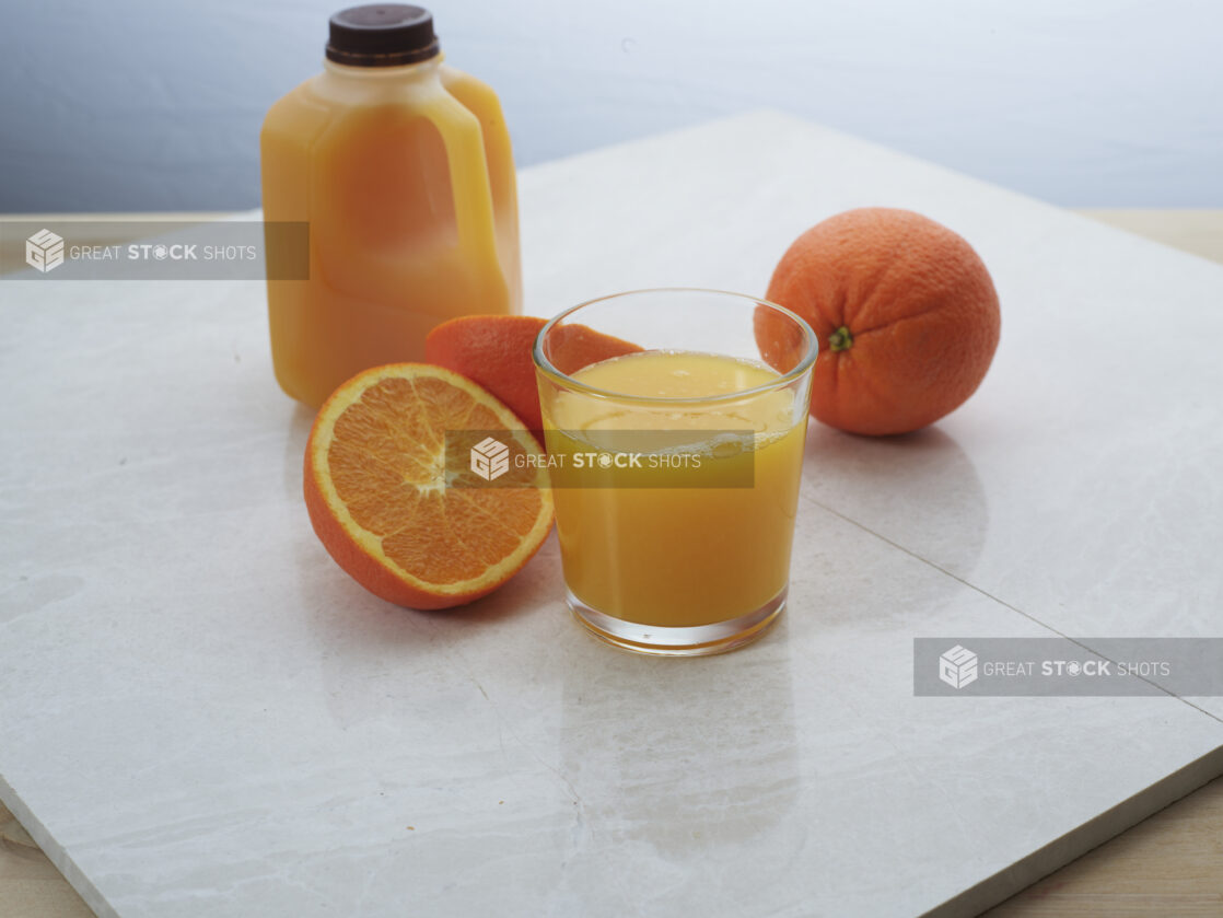 Small glass of freshly-squeezed orange juice with whole and halved oranges and juice jug in background