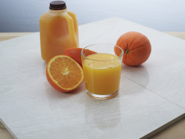 Small glass of freshly-squeezed orange juice with whole and halved oranges and juice jug in background