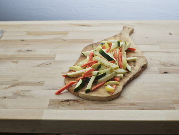 Sliced zucchini, carrots, and summer squash on a natural wood paddle, close-up