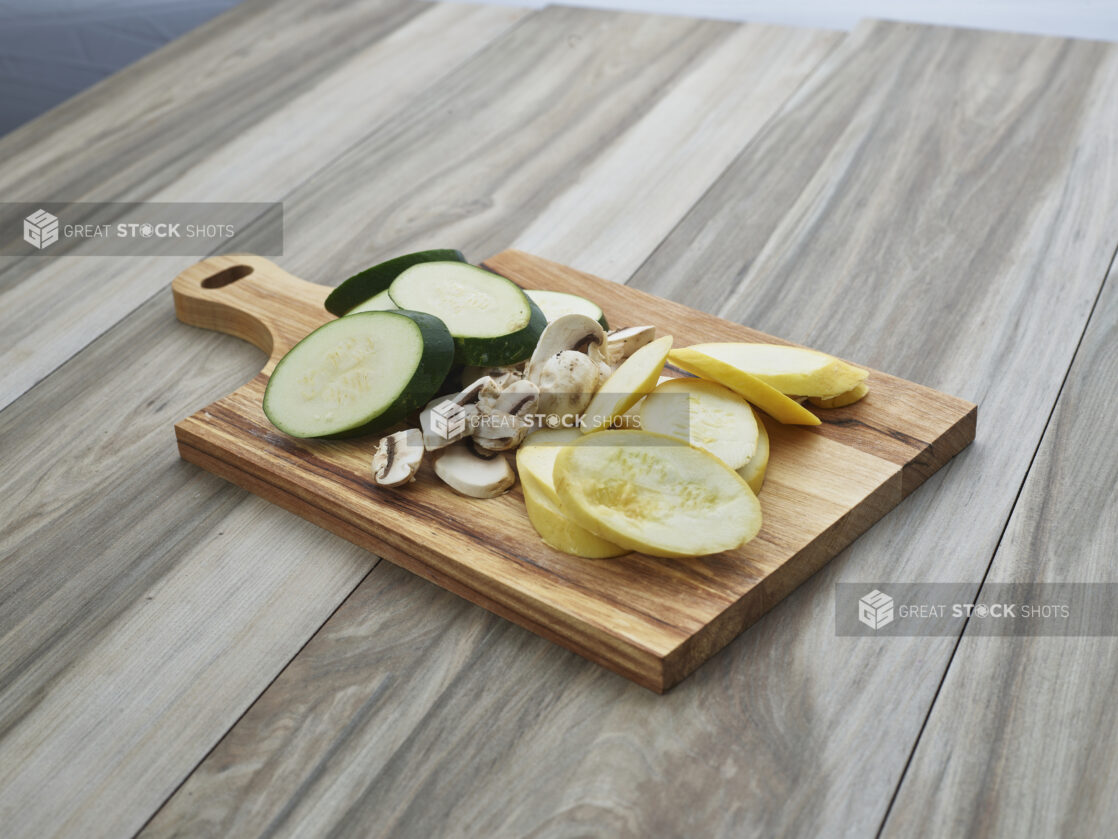 Sliced zucchini, summer squash, and mushrooms arranged on a wood paddle, close-up