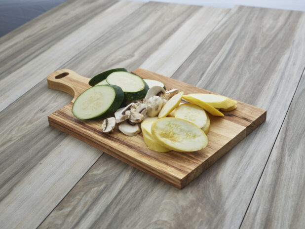 Sliced zucchini, summer squash, and mushrooms arranged on a wood paddle, close-up
