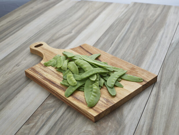 Fresh snow pea pods on a rectangular wood paddle, close-up