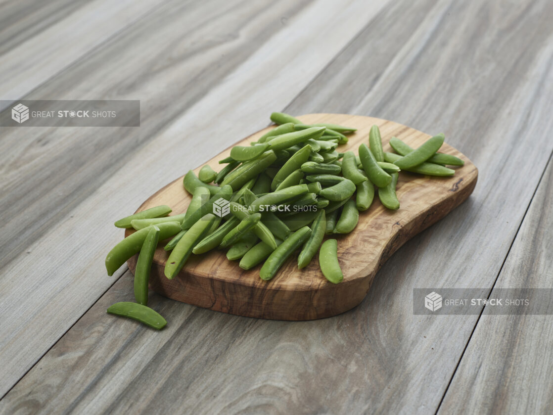 Fresh snap peas piled on a rounded natural wood board, close-up