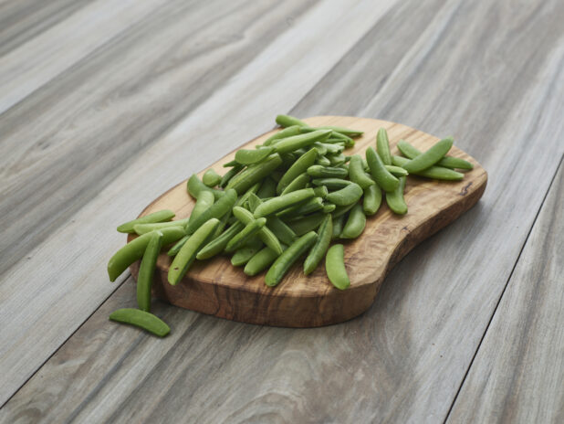 Fresh snap peas piled on a rounded natural wood board, close-up