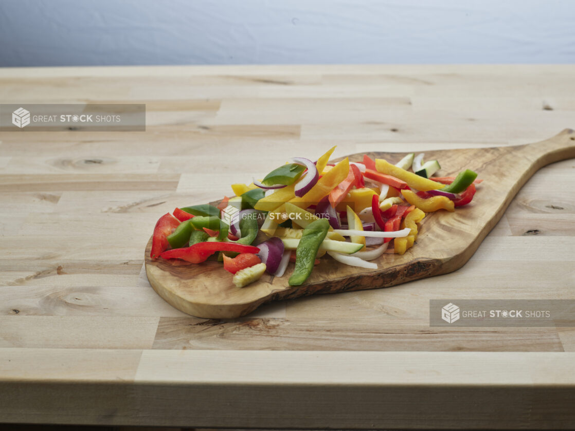 Sliced bell peppers, zucchini, summer squash, and onions on a natural wood paddle, close-up