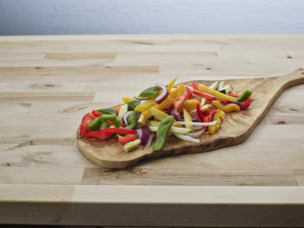 Sliced bell peppers, zucchini, summer squash, and onions on a natural wood paddle, close-up