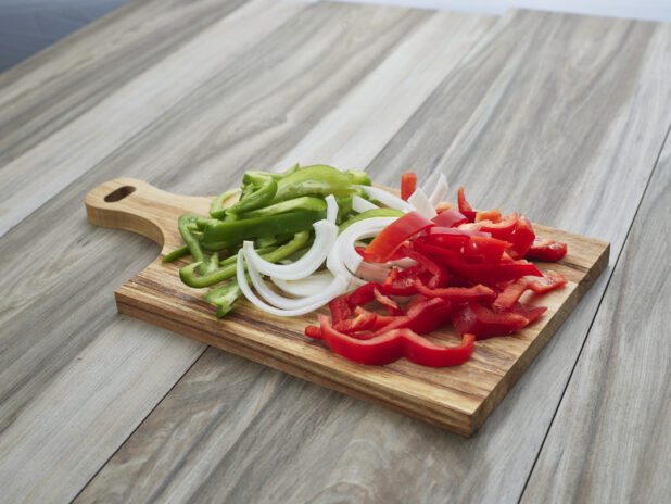 Sliced raw green and red peppers and white onions on a wooden paddle, close-up