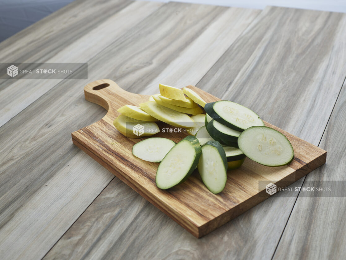 Slices of raw zucchini and summer squash in separate piles on a wooden paddle, close-up